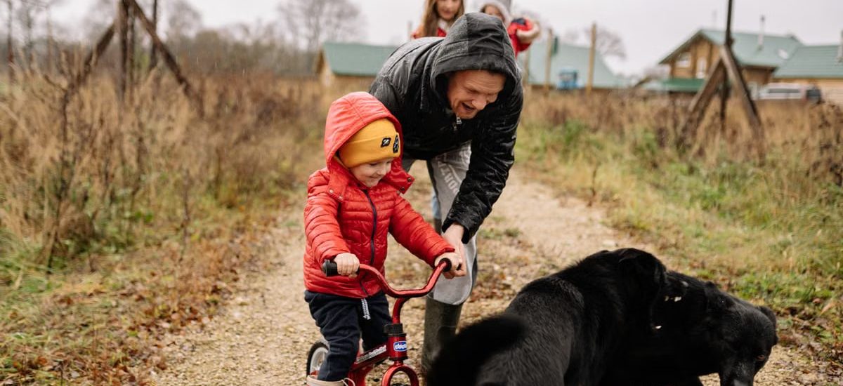 Dad helps son ride a bike on a path with family and dog, capturing a joyful outdoor moment.