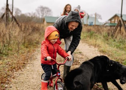 Dad helps son ride a bike on a path with family and dog, capturing a joyful outdoor moment.