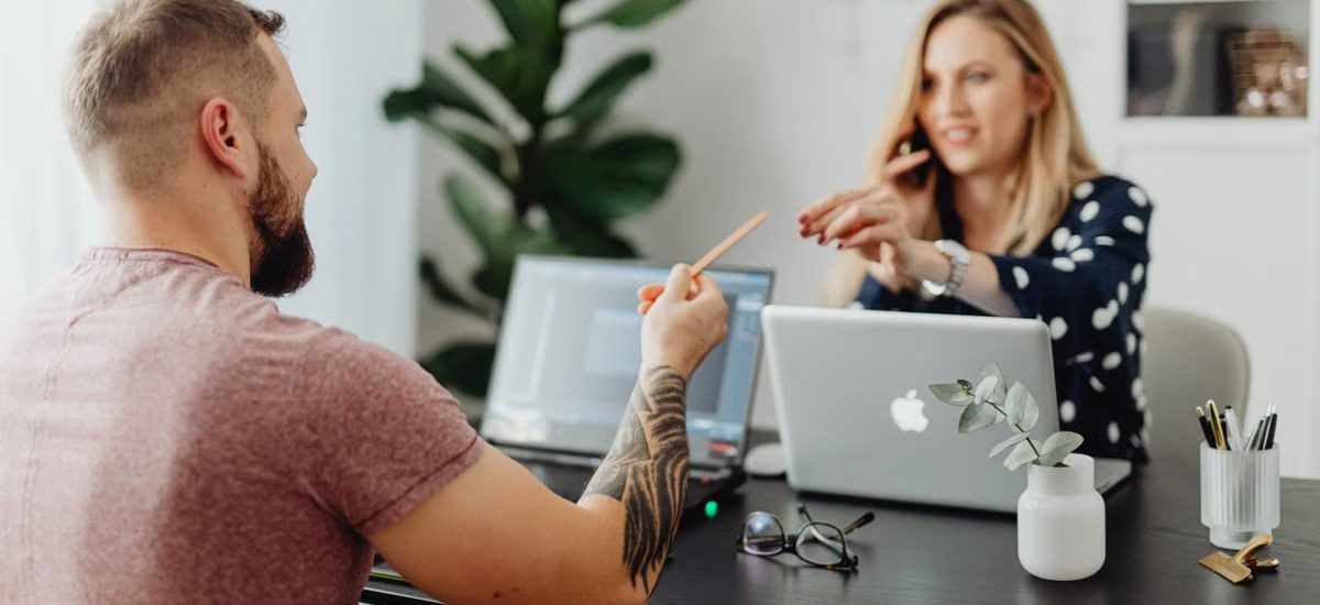 Man and woman discussing work at a table with laptops in a modern office.